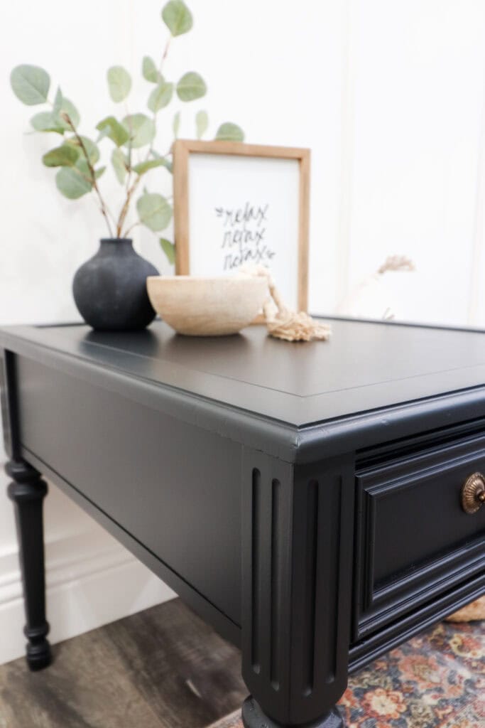 Black side table with a drawer, decorated with a vase, bowl, and framed art on top.