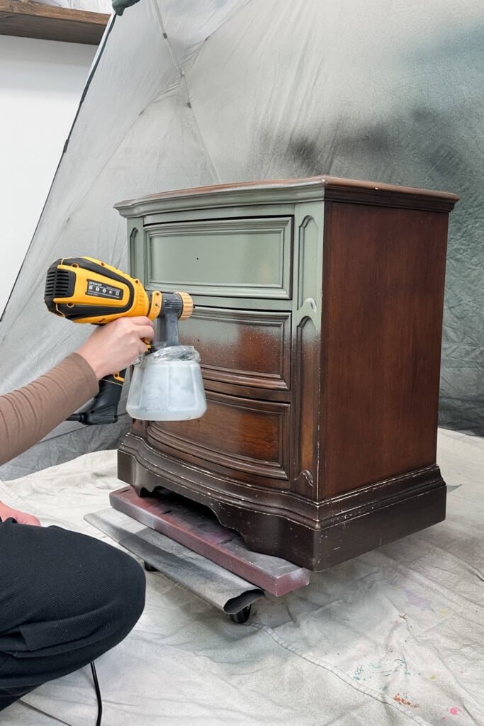 Person spray painting a wooden dresser green, with protective sheet in the background.