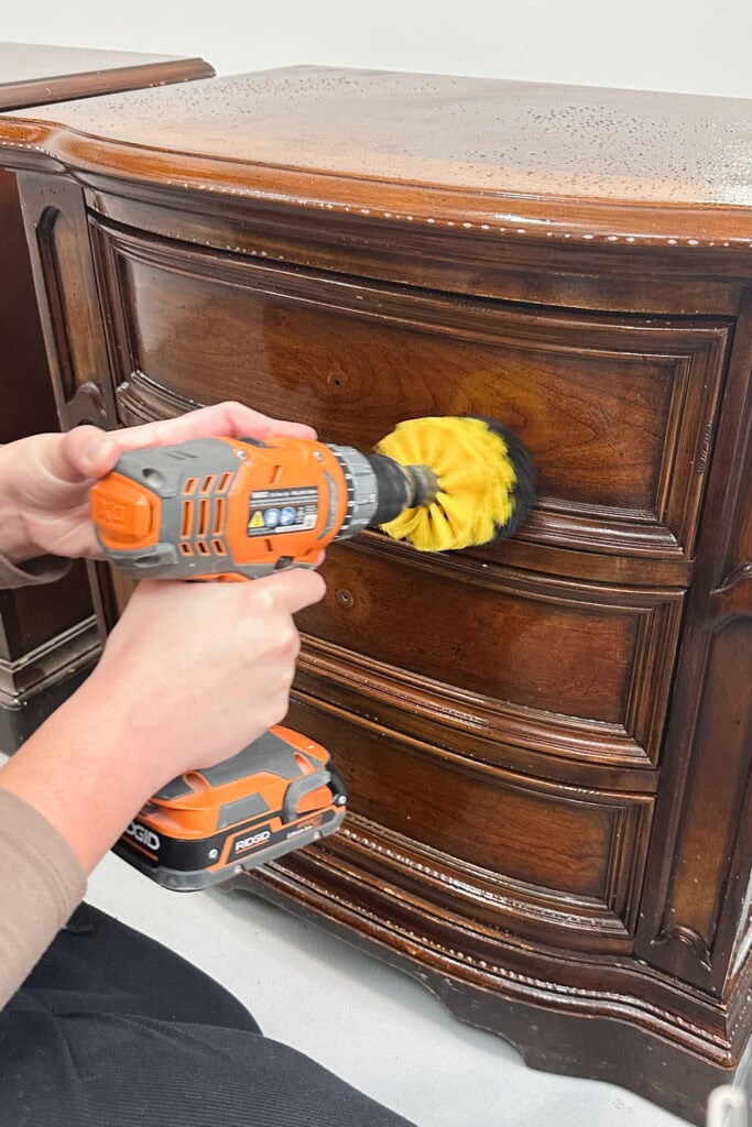 Person using a power drill with a brush attachment to clean or polish a wooden dresser drawer.