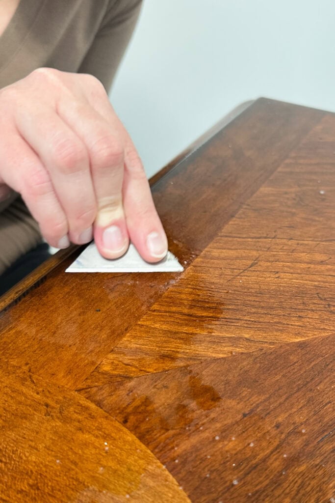 A person scraping the surface of a wooden table with a razor blade