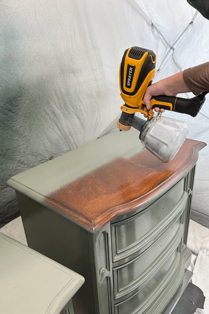 A hand uses a paint sprayer to apply paint to the top of a wooden dresser