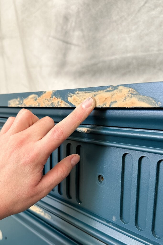 A hand spreads wood filler on the edge of a blue wooden surface.