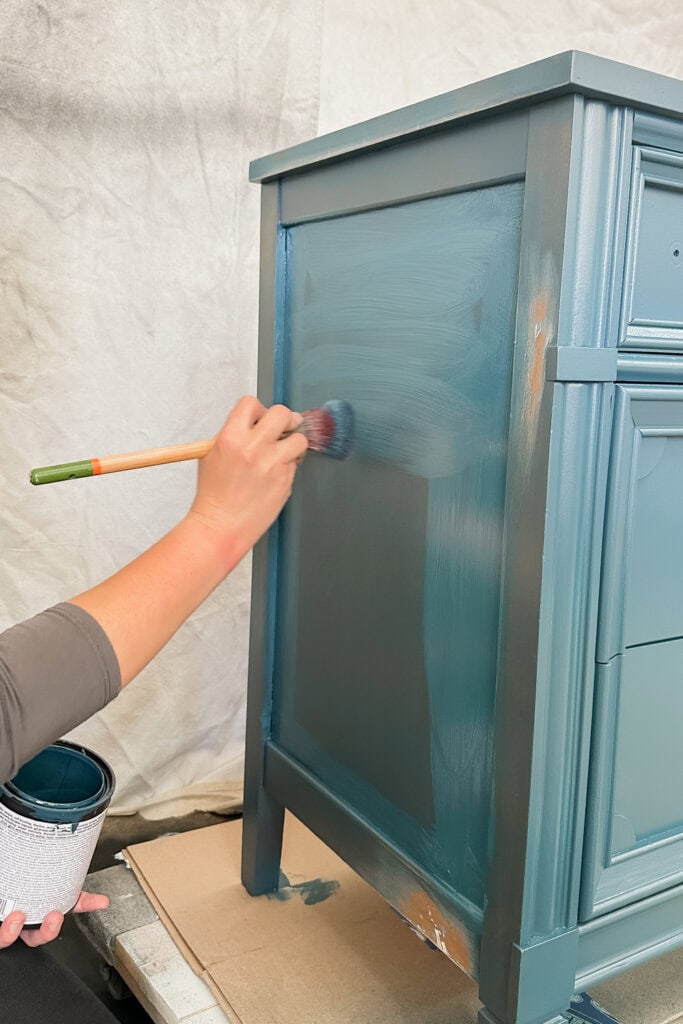 A person painting a blue wooden dresser with a brush, holding a paint can in their other hand.