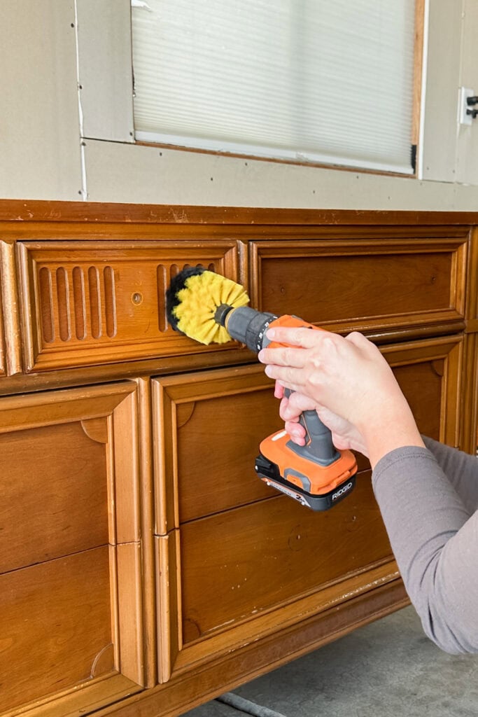 A person uses a power drill with a brush attachment to clean a wooden cabinet drawer.