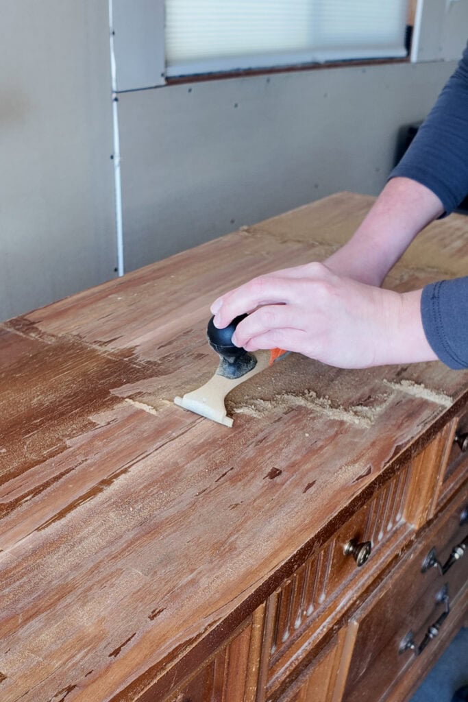 A person scrapes old finish off a wooden dresser with carbide scraper