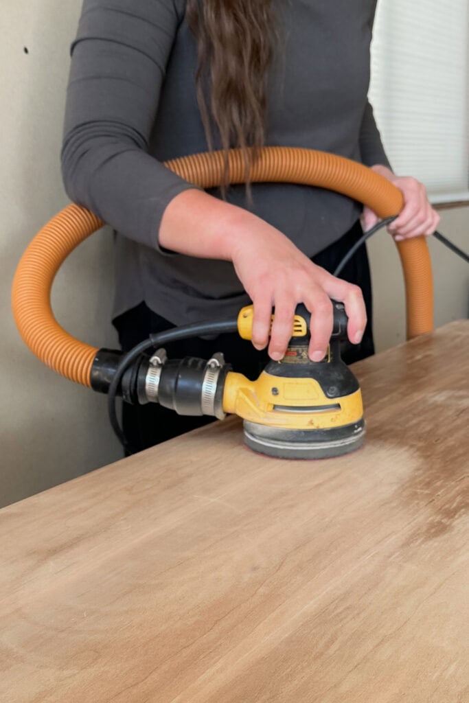 Person using a orbital sander to smooth a wooden surface, holding a vacuum hose for dust.