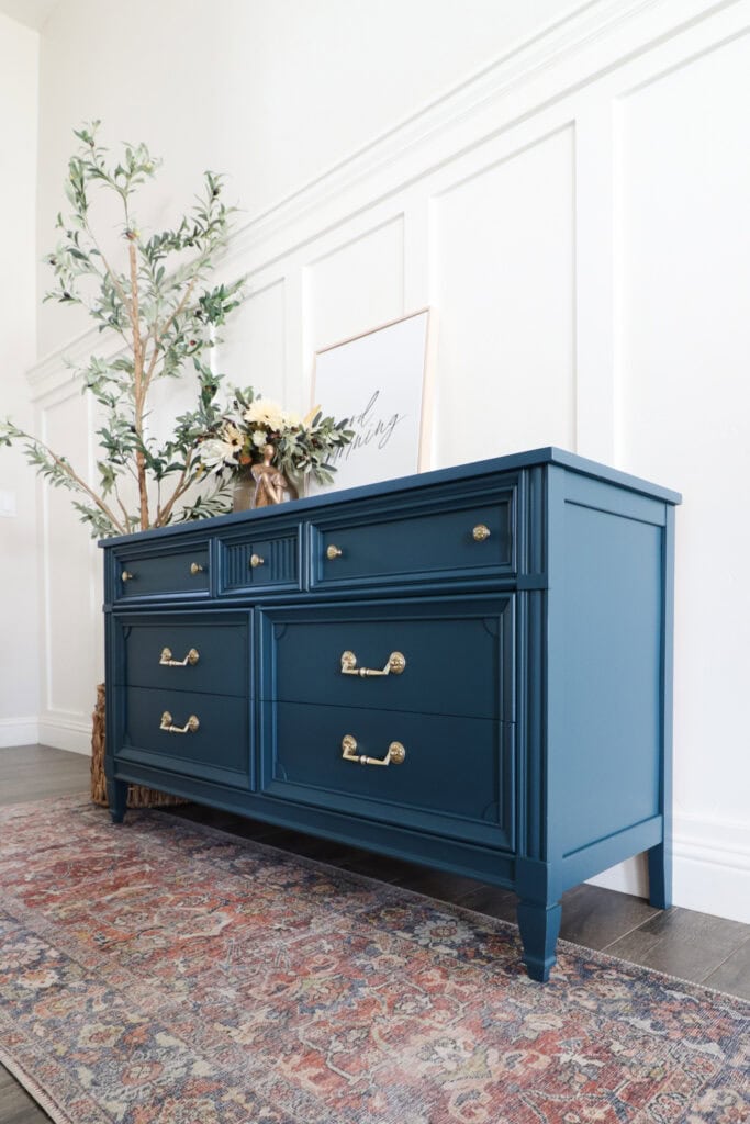 A blue dresser with gold handles sits against a white wall, topped with decor and a leafy plant nearby.
