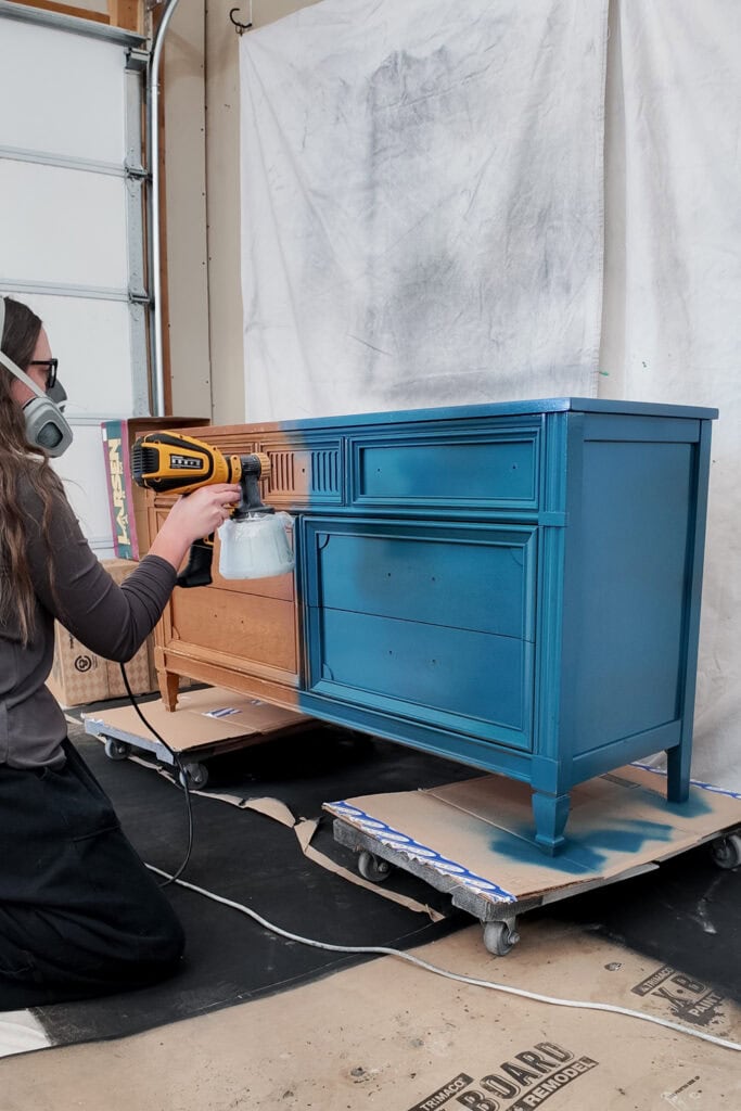 Natalie wearing a respirator spray paints a wooden dresser blue