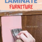 A hand painting pink chalk paint on a brown laminate furniture surface with a brush.