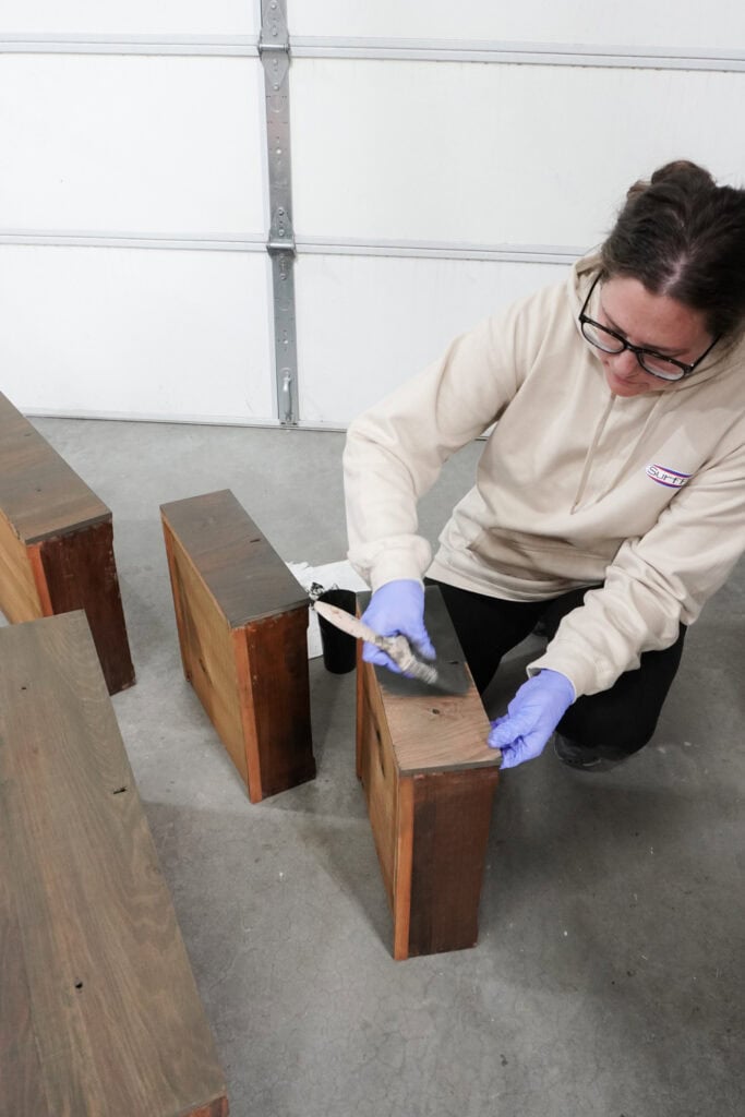 A person wearing gloves applies paint wash to a wooden drawer in a garage or workshop.