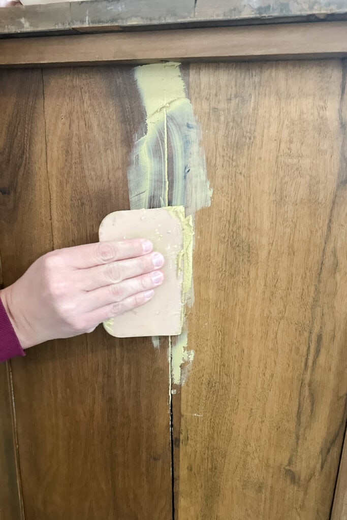 A hand uses a putty knife to apply wood filler on a crack in wood surface