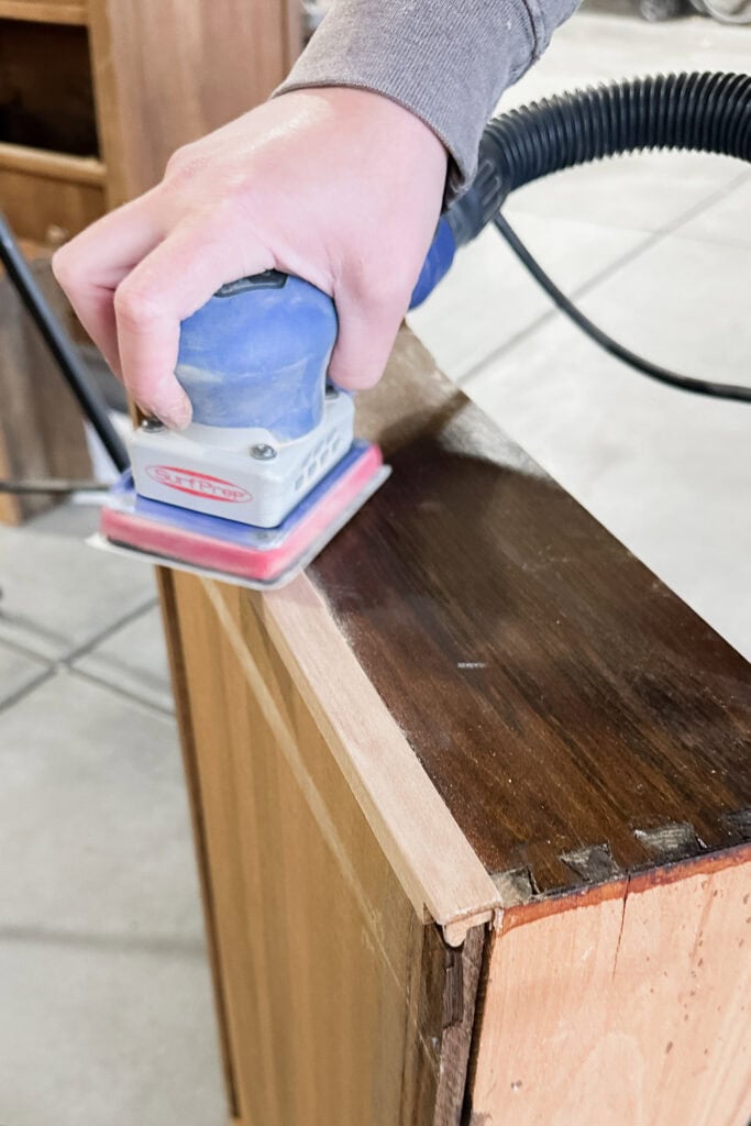 A person uses an electric sander to refinish the edge of a wooden drawer