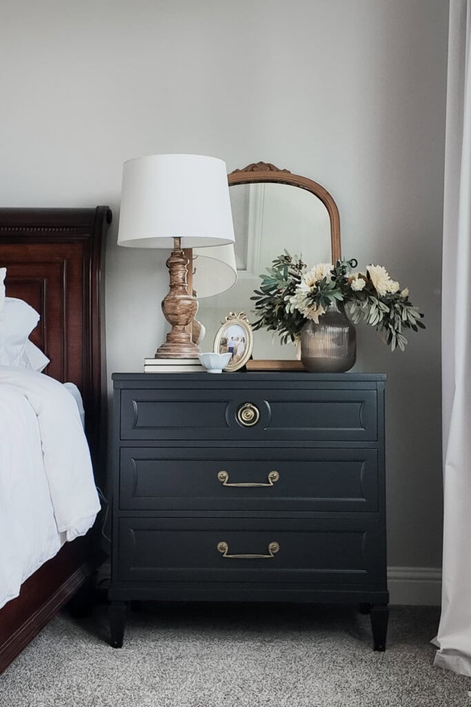 Dark nightstand with lamp, framed photo, flowers in vase, and mirror beside a bed with white bedding.