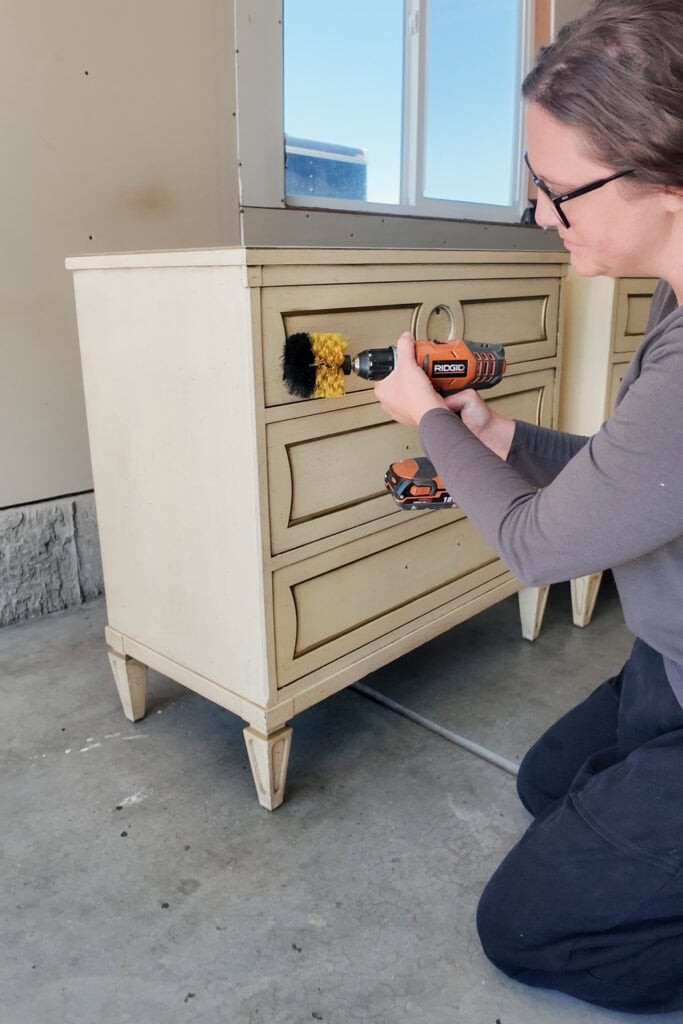 Natalie using a power drill with a brush attachment to clean a drawer front on a cream dresser.