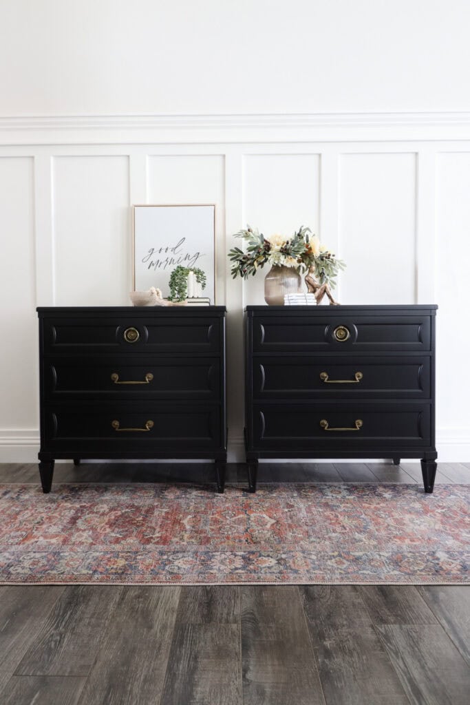Two black nightstands with gold handles, floral decor, and framed art, on a rug against a white paneled wall.