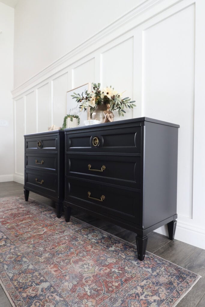 Two black nightstands with gold handles, floral decor on top, on a patterned rug against a white paneled wall.