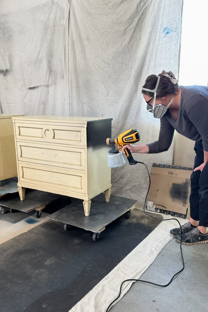 Person wearing a mask spray-painting a light-colored dresser black in a protected work area.