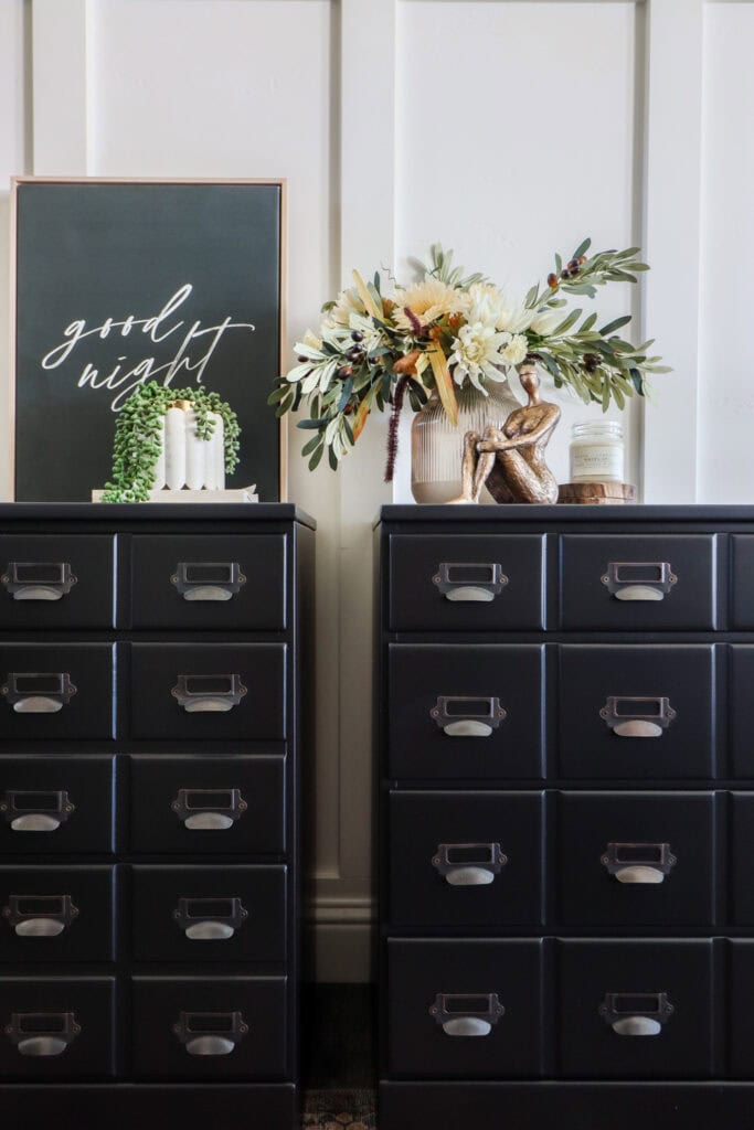 Closeup of two black nightstands with card catalog pulls, decor items, flowers, and a framed good night sign on top.