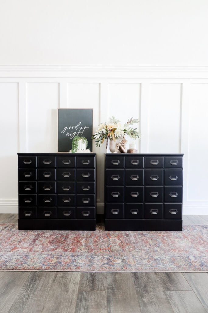Two black nighstands with card catalog pulls, a framed sign, and flowers on top, set against a white paneled wall.
