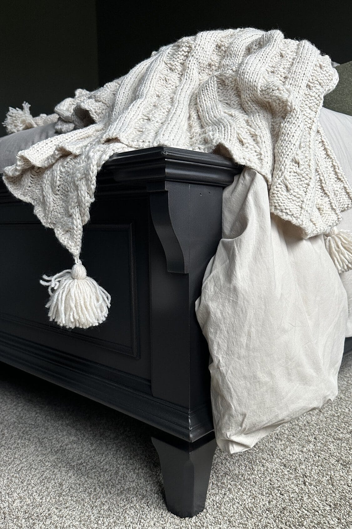 Cream knit blanket with tassel draped over the dark footboard of a wooden updated bed in a cozy bedroom.