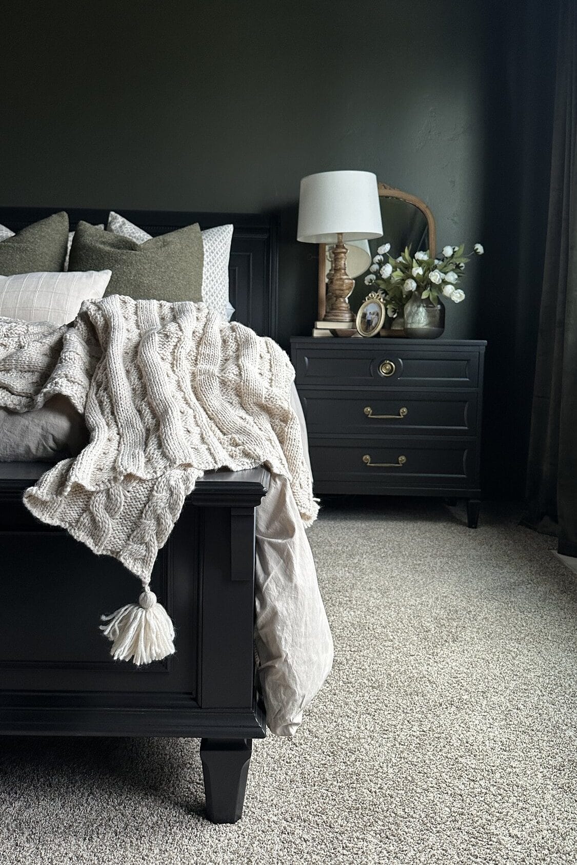 Cozy bedroom with black updated bed, knit blanket, nightstand, lamp, and flowers on beige carpet.