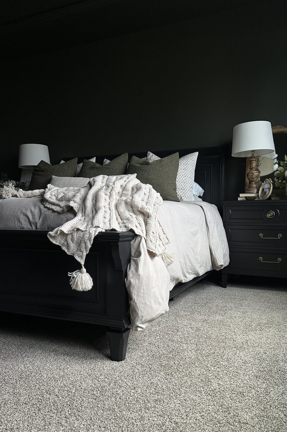 A cozy DIY black painted bed with beige bedding, dark green pillows, and two bedside tables with lamps on a carpeted floor.