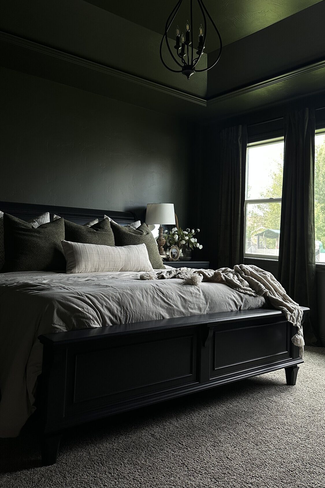 A dark green modern bedroom with a diy painted black bed, tan bedding, lamps, and large window with green velvet curtains.