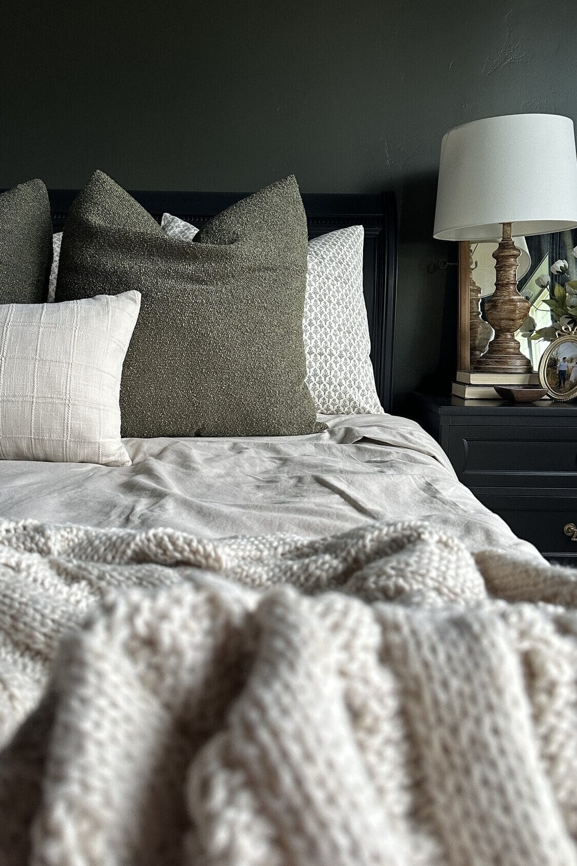 Cozy painted bed with textured pillows, blanket, and a lamp on a dark nightstand with a clock and decor.