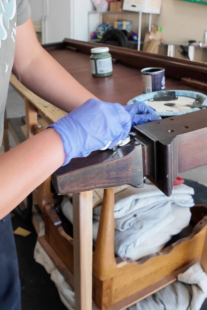 Person wearing blue gloves stains wood furniture with a cloth in a garage.