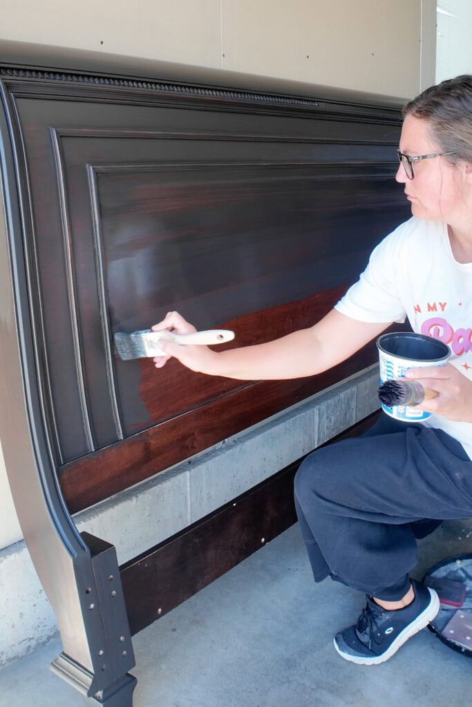 A woman paints a wooden headboard with a brush while holding a can of paint