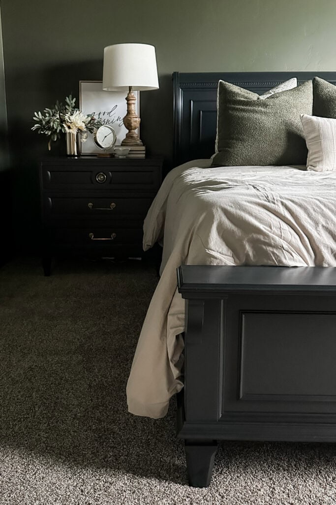 Dark bed frame with green pillows, beige bedding, and a nightstand with a lamp, clock, and decor on carpeted floor.