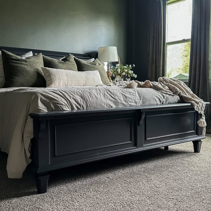 A dark wood bed with neutral bedding, pillows, and a lamp sits by a window in a cozy, carpeted room.