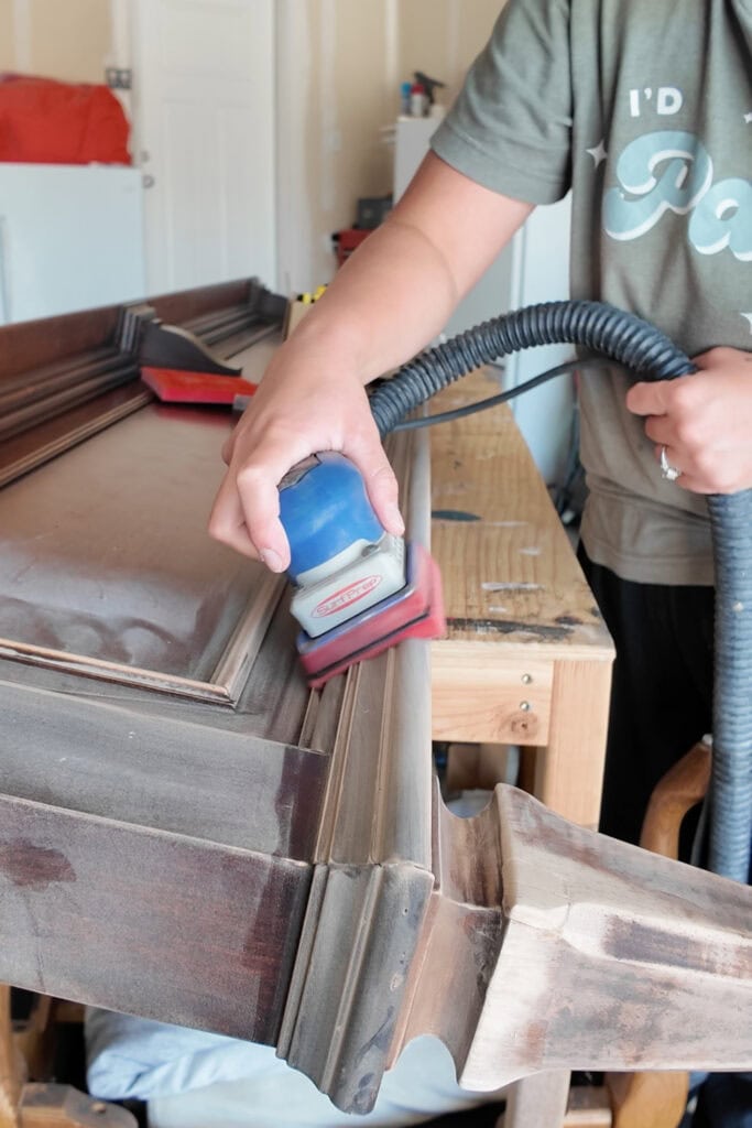 Person sanding a wooden furniture piece with a power sander and vacuum in a workshop.