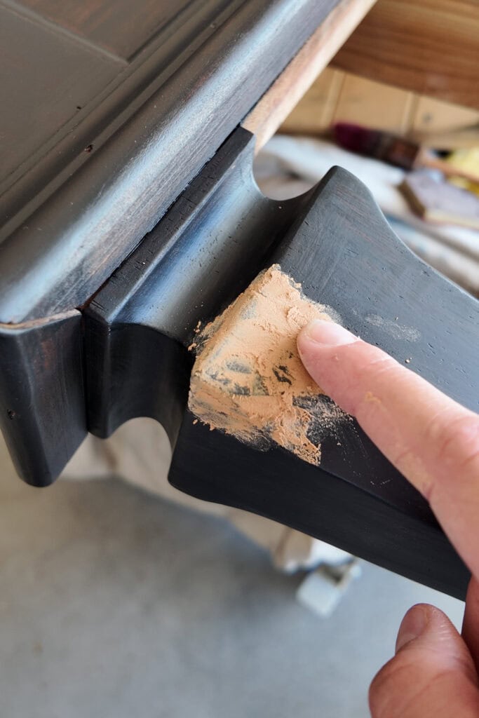 A hand applies wood filler to a chipped area on a dark colored wooden furniture piece.