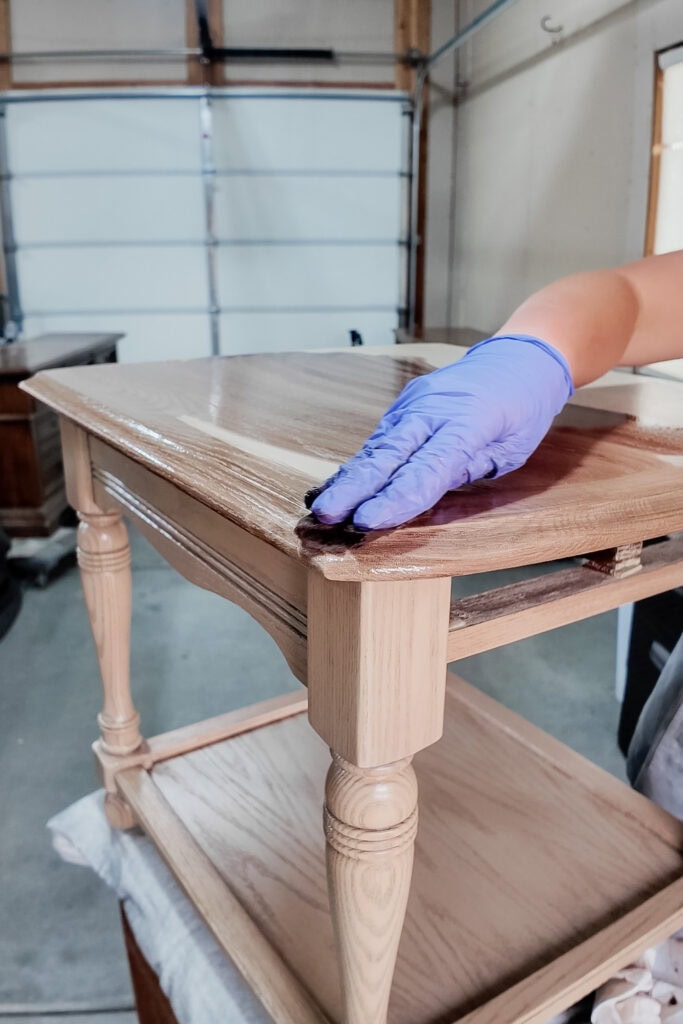 A gloved hand applies stain to a wooden side table in a garage workshop.