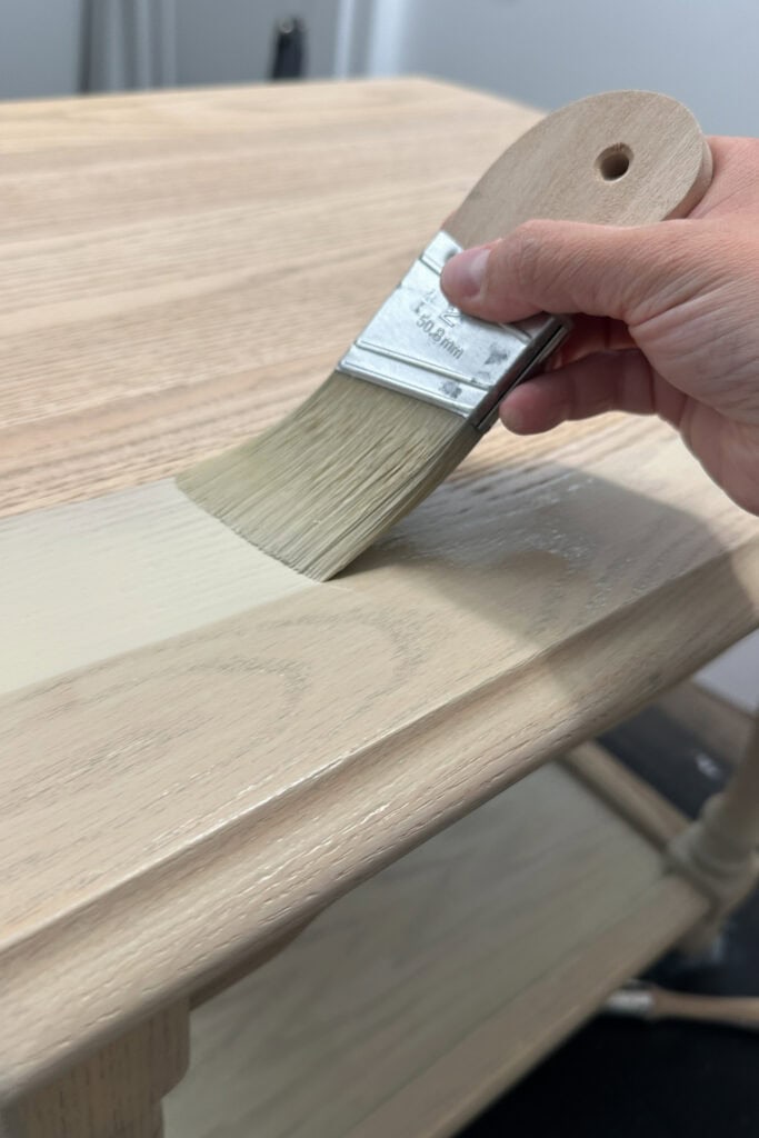 A hand paints a wooden table with a brush, applying a light colored paint wash to the surface.