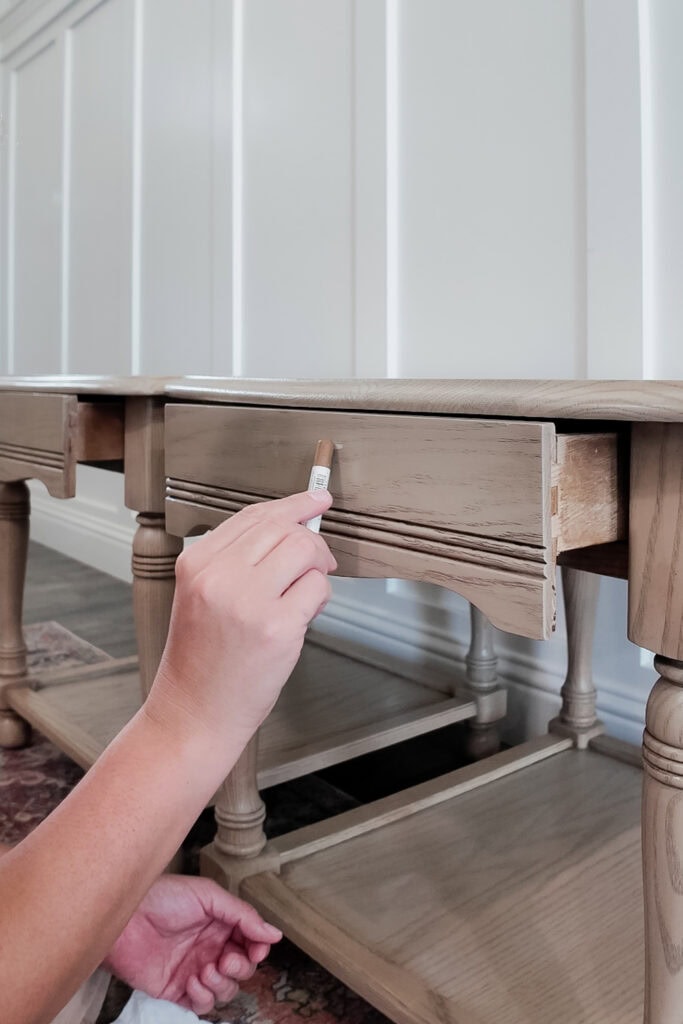 A hand uses a wood stain marker to touch up a wooden drawer on a light colored table.