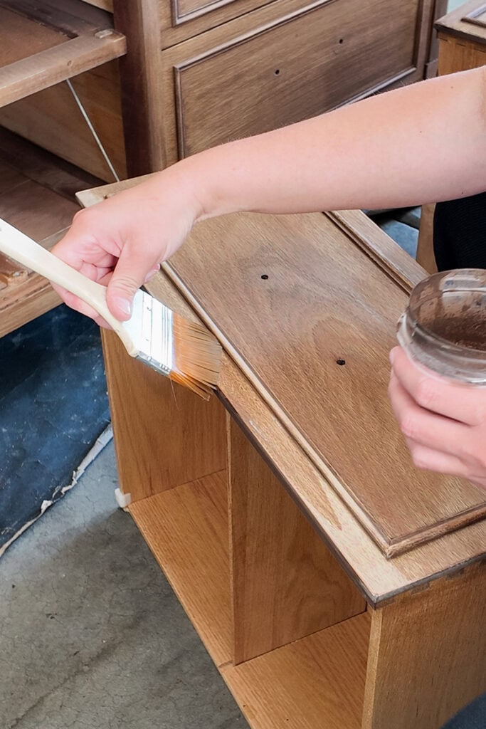 A person paints brown paint wash on a wooden drawer front using a paintbrush.