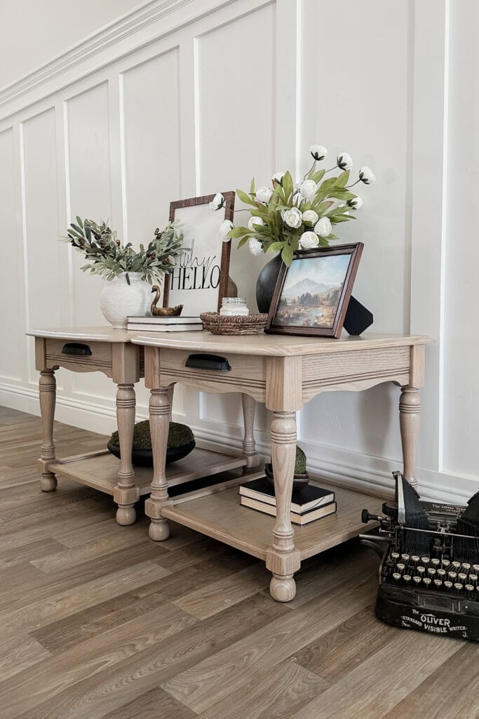 Two wooden tables with decor, flowers, books, a framed photo, and a vintage typewriter on a wooden floor.