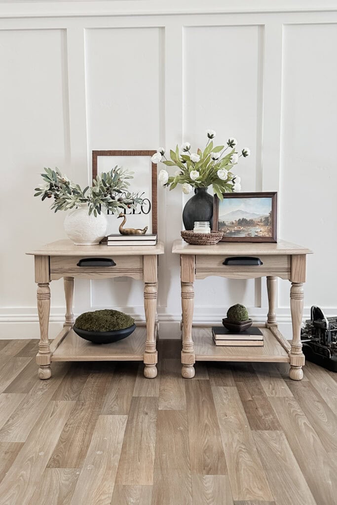 Two wooden side tables with plants, books, framed art, and decor on a wood floor against a white paneled wall.