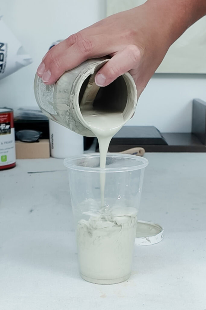 A hand pours light green paint from a jar into a clear plastic cup with water