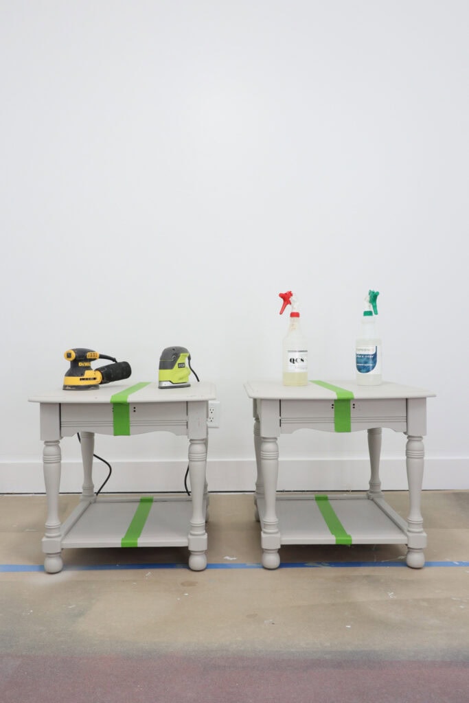 Two gray end tables with tape, each holding tools and cleaners, against a white wall on a painted floor.