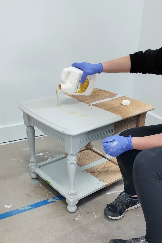 A person pours chemical stripper from a bottle onto a small table, wearing blue gloves and black clothes.