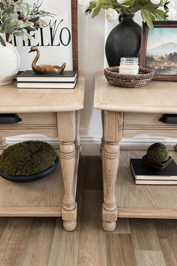 Two wooden side tables with decor, books, greenery, and art on top and shelves, on a wood floor.