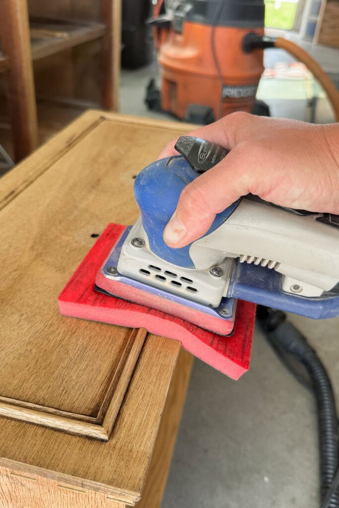 A hand holds a power sander with an oversized foam pad, sanding a wooden surface.