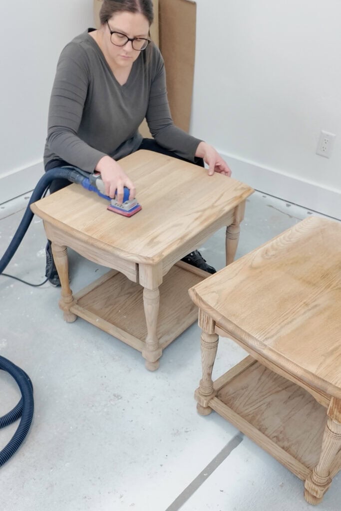 Woman sanding a wooden side table with surfprep sander, with another table beside her on the floor.