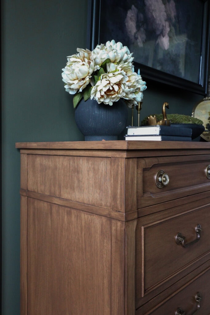 Wooden dresser with gold handles, a vase of white flowers, and decor items on top against a dark green wall.