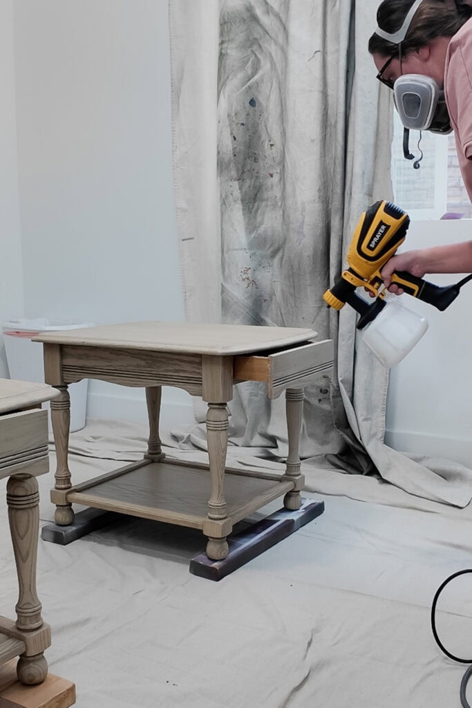 Person wearing a respirator uses a paint sprayer to spray topcoat on a wooden side table indoors.
