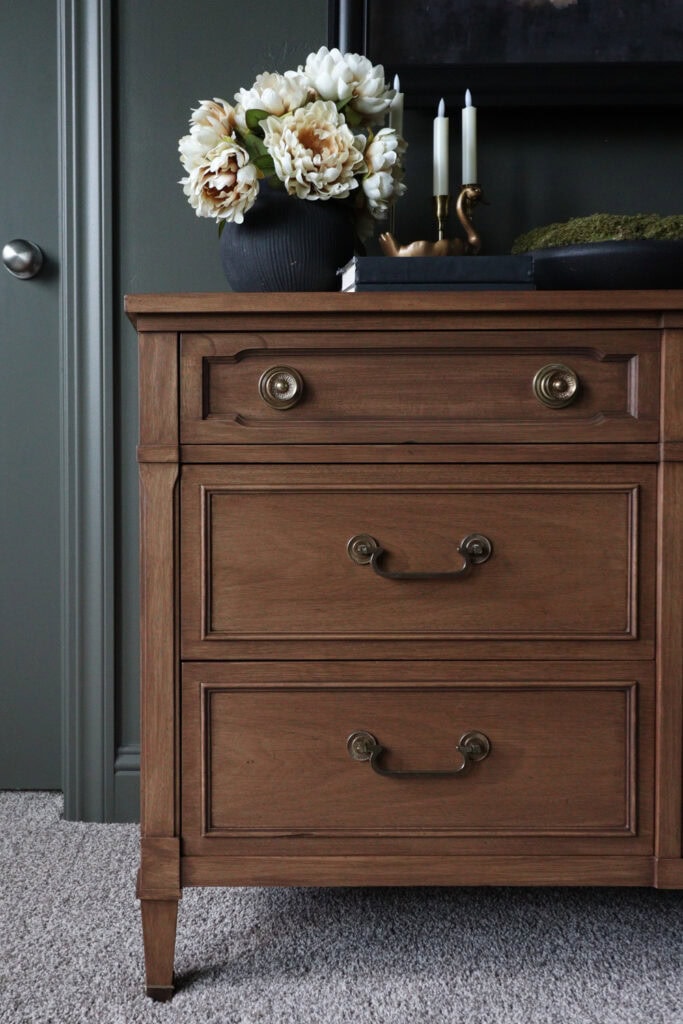 Wooden dresser with gold handles, topped with white flowers in a vase, candles, and a decorative tray.