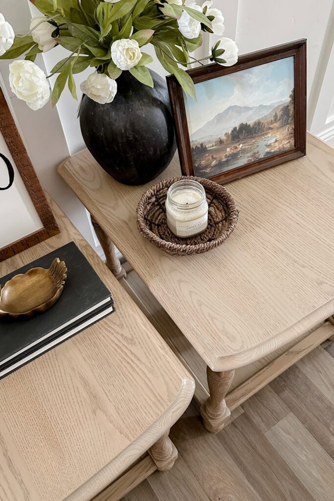 Two light wood tables with a vase of white flowers, framed art, candle, books, and brass decor displayed.
